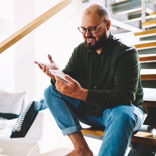Mann sitzt mit einem Smartphone auf einer modernen Mittelholmtreppe und lächelt, während er auf das Display sieht