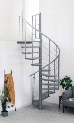 Spindeltreppe Spino Smart in Grau aus Metall im modernen Wohnraum. Die gewendelte Treppe führt nach oben, das Geländer besteht aus senkrechten Streben und Edelstahlseilen. Im Hintergrund stehen ein Sofa, Pflanzen und Deko.