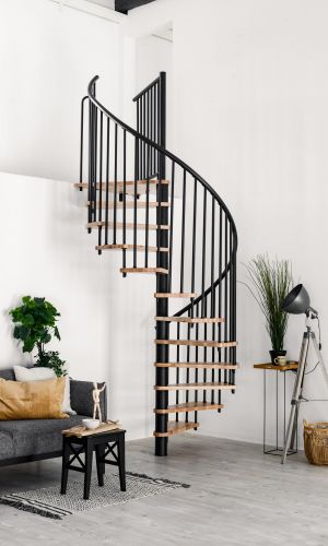 Gesamtaufnahme der Spindeltreppe Spiral Smart mit Buchenstufen und schwarzem Geländer, montiert in einem weißen Raum. Links steht ein graues Sofa mit weiß-braunen Kissen und ein schwarzer Beistelltisch mit Deko. Rechts steht eine graue Stehlampe, eine Zimmerpflanze und ein Bastkorb.