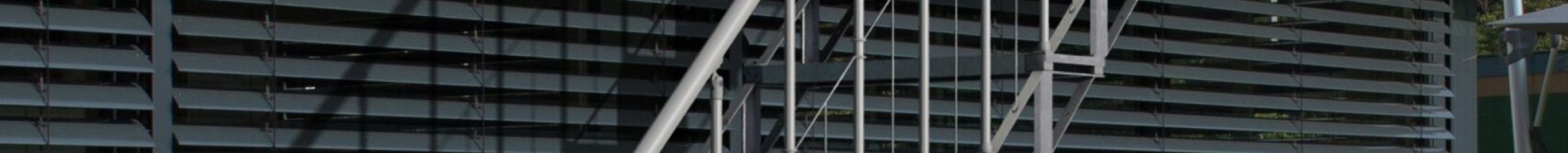 Außentreppe Sun Step aus verzinktem Stahl an einer modernen Hausfassade. Die Treppe hat 12 einzelne Stufen mit Gitterrost-Auflage, ein beidseitiges Geländer mit senkrechten Stäben und einem Handlauf aus Edelstahl. Zugang von der Terrasse in den Garten.