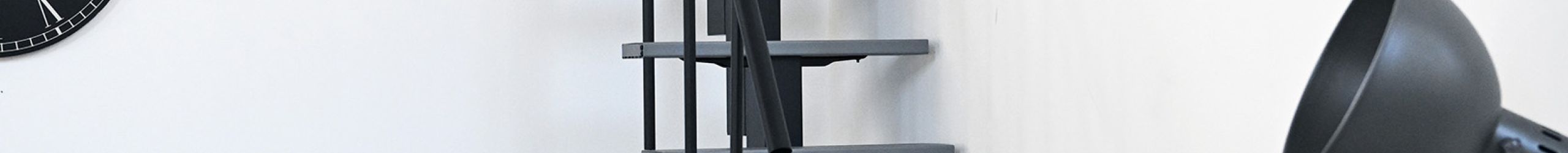 Raumspartreppe mit versetzt angeordneten grauen Stufen und schwarzer Mittelwange in modernem Wohnraum mit hellem Holzboden. Die Treppe ist einseitig mit einem schwarzen Handlauf versehen. Links steht ein graues Sofa, rechts eine schwarze Stehlampe neben einer grünen Pflanze im Drahtkorb.