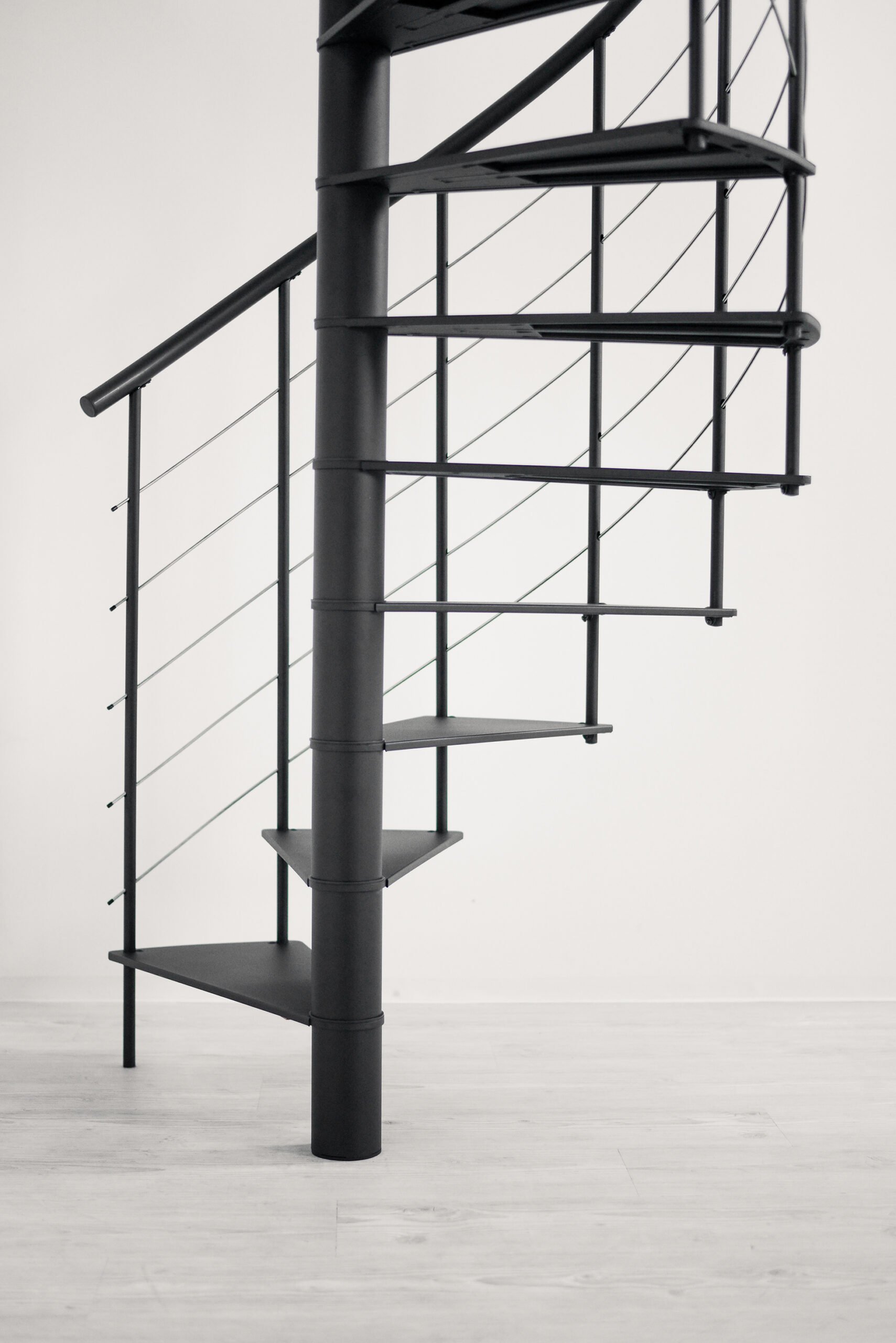 Seitenansicht der modernen Spindeltreppe „Milano“ aus schwarzem Metall mit offenen Trittstufen, zentrales Rundrohr als tragendes Element und einem Geländer aus vertikalen Metallstreben sowie horizontalen Spannseilen.