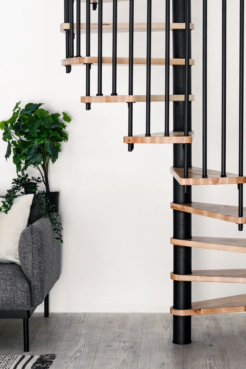 Spindeltreppe Spiral Wood mit schwarzen Metallstäben und Buchenstufen, die spiralförmig um eine schwarze Mittelsäule führen. Daneben ein graues Sofa mit weißen Kissen und eine grüne Zimmerpflanze. Der Raum ist hell und minimalistisch gestaltet.