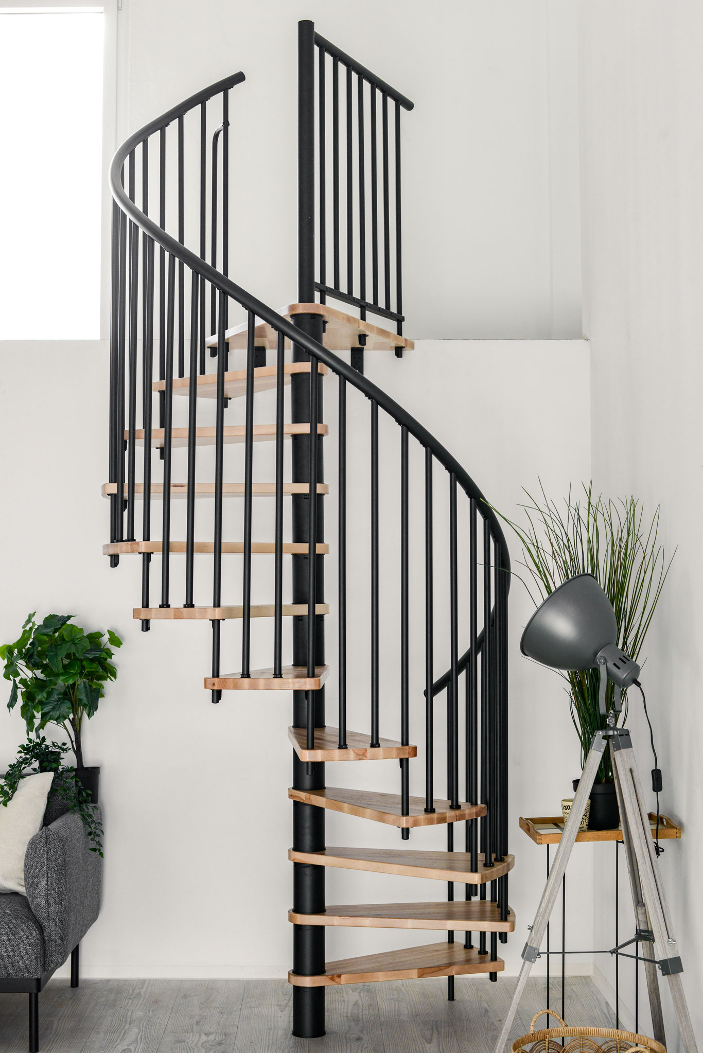 Spindeltreppe Spiral Smart mit schwarzem Metallgeländer mit PVC-Handlauf und Buchenstufen, die spiralförmig um eine schwarze Mittelsäule führen. Links daneben ein graues Sofa mit weißem Kissen und einer Pflanze. Rechts daneben eine Pflanze und eine graue Stehlampe.