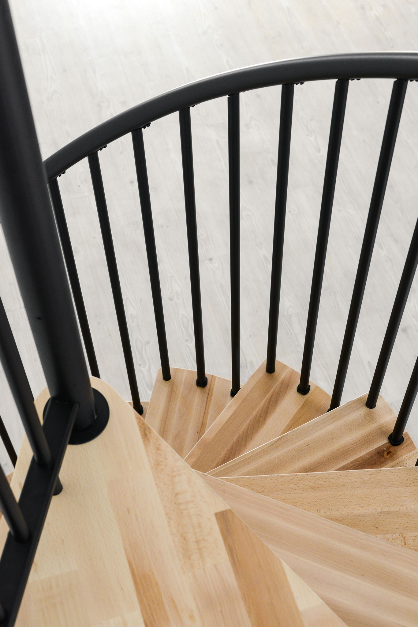 Nahaufnahme der hellen Buchenstufen der Spiral Smart von oben. Außen an den Stufen ist ein schwarzes Metallgeländer mit vertikalen Stehern befestigt, die oben am schwarzem PVC-Handlauf befestigt sind.