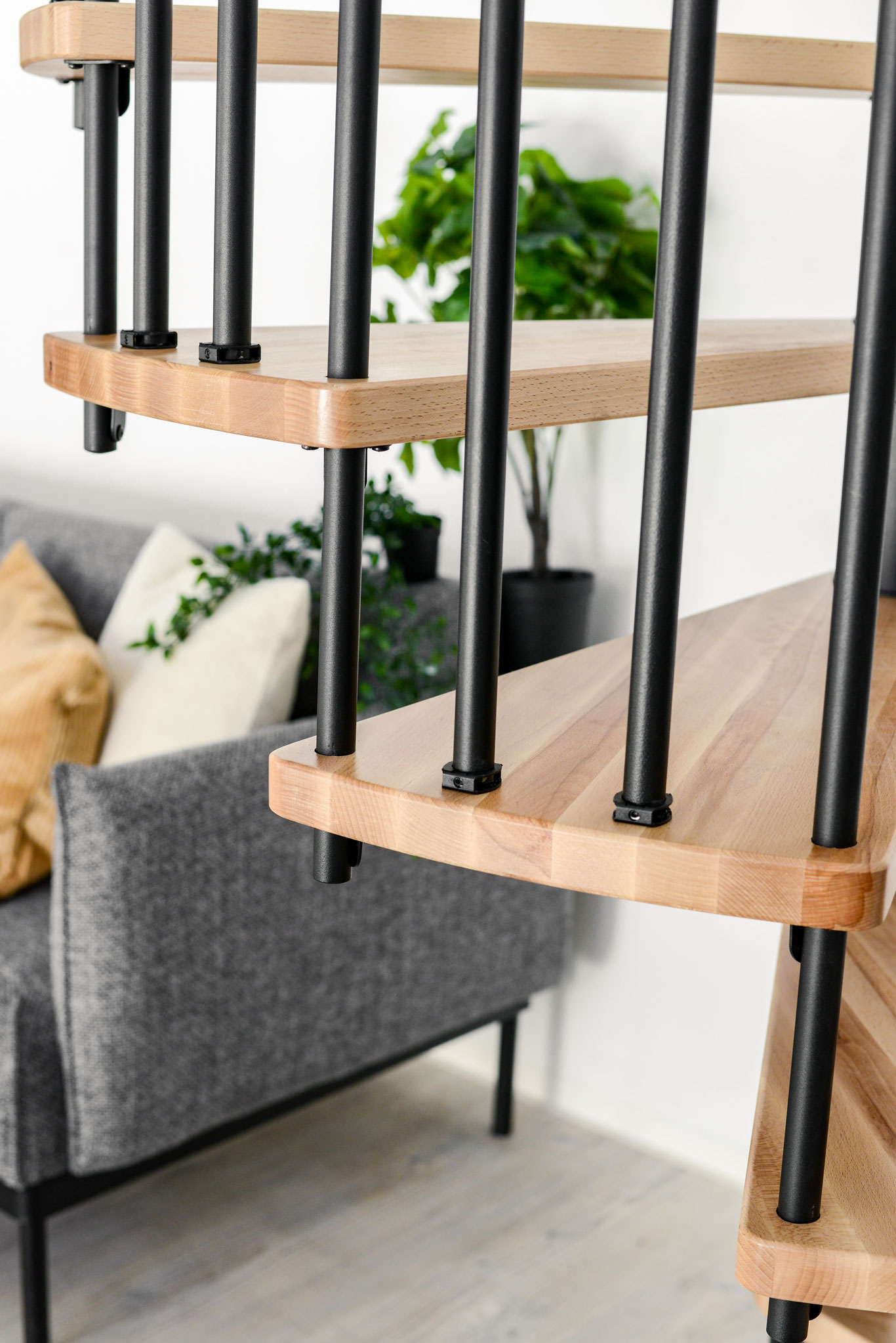 Nahaufnahme der Spindeltreppe Spiral Smart mit Buchenstufen und schwarzen Geländerstäbe von der Seite. Im Hintergrund steht eine graue Couch mit Pflanzen und Polstern.