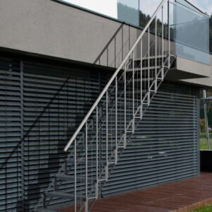 Außentreppe Sun Step aus verzinktem Stahl an einer modernen Hausfassade. Die Treppe hat 12 einzelne Stufen mit Gitterrost-Auflage, ein beidseitiges Geländer mit senkrechten Stäben und einem Handlauf aus Edelstahl. Zugang von der Terrasse in den Garten.