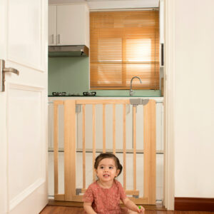 Kinderschutztür Britt in Türrahmen montiert, dahinter eine Küche. Ein kleines Kind spielt sicher im Vordergrund auf dem Holzboden mit buntem Spielzeug. Die Tür besteht aus hellem Holz mit vertikalen Streben und grauen Schließelementen.