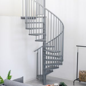 Graue Spindeltreppe Rondo Smart aus Metall mit runden Stufen und senkrechten Geländerstäben. Die platzsparende Treppe führt spiralförmig nach oben und steht in einem modernen Wohnraum mit Sofa, Pflanzen und Kleiderständer.