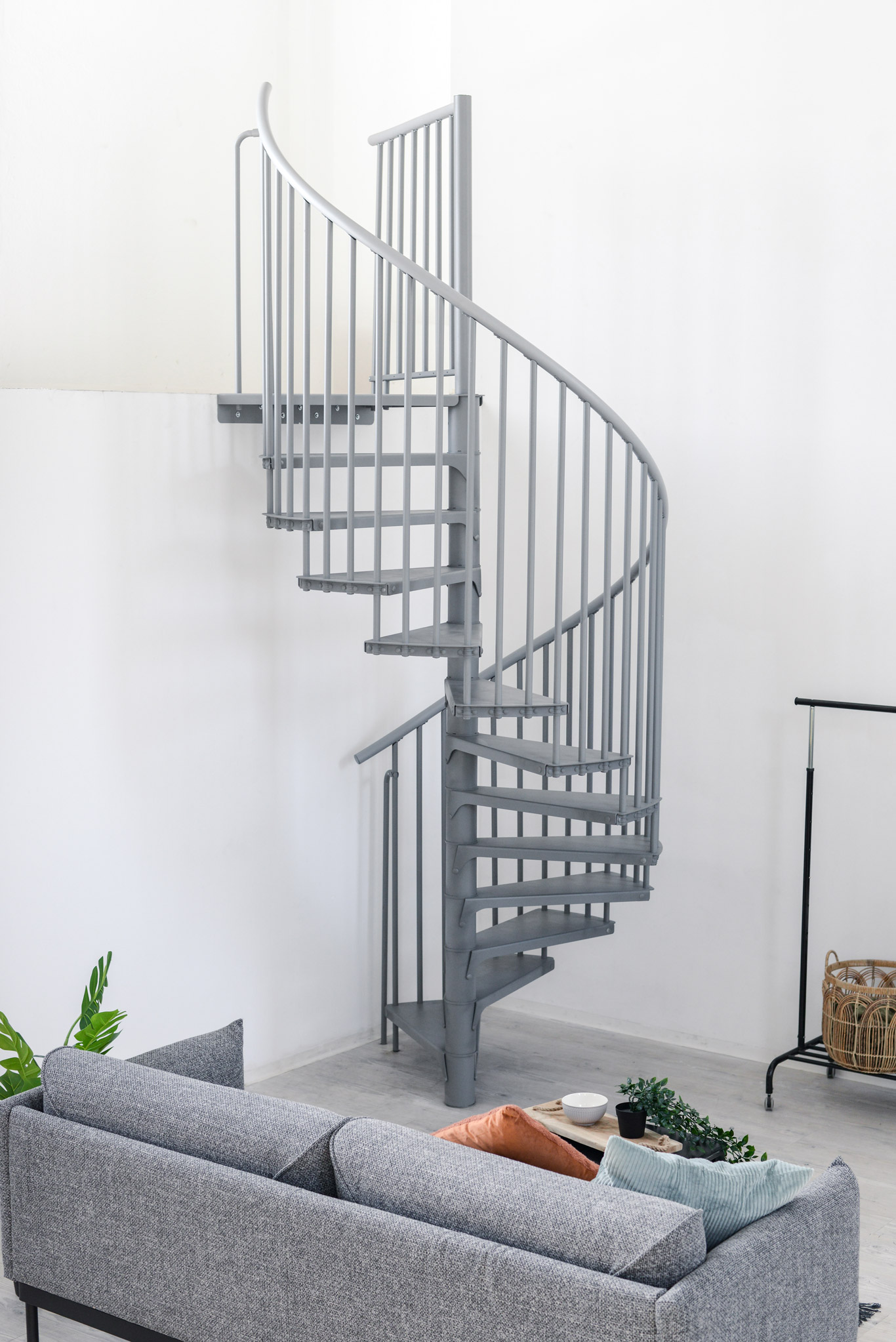 Graue Spindeltreppe Rondo Smart aus Metall mit runden Stufen und senkrechten Geländerstäben. Die platzsparende Treppe führt spiralförmig nach oben und steht in einem modernen Wohnraum mit Sofa, Pflanzen und Kleiderständer.
