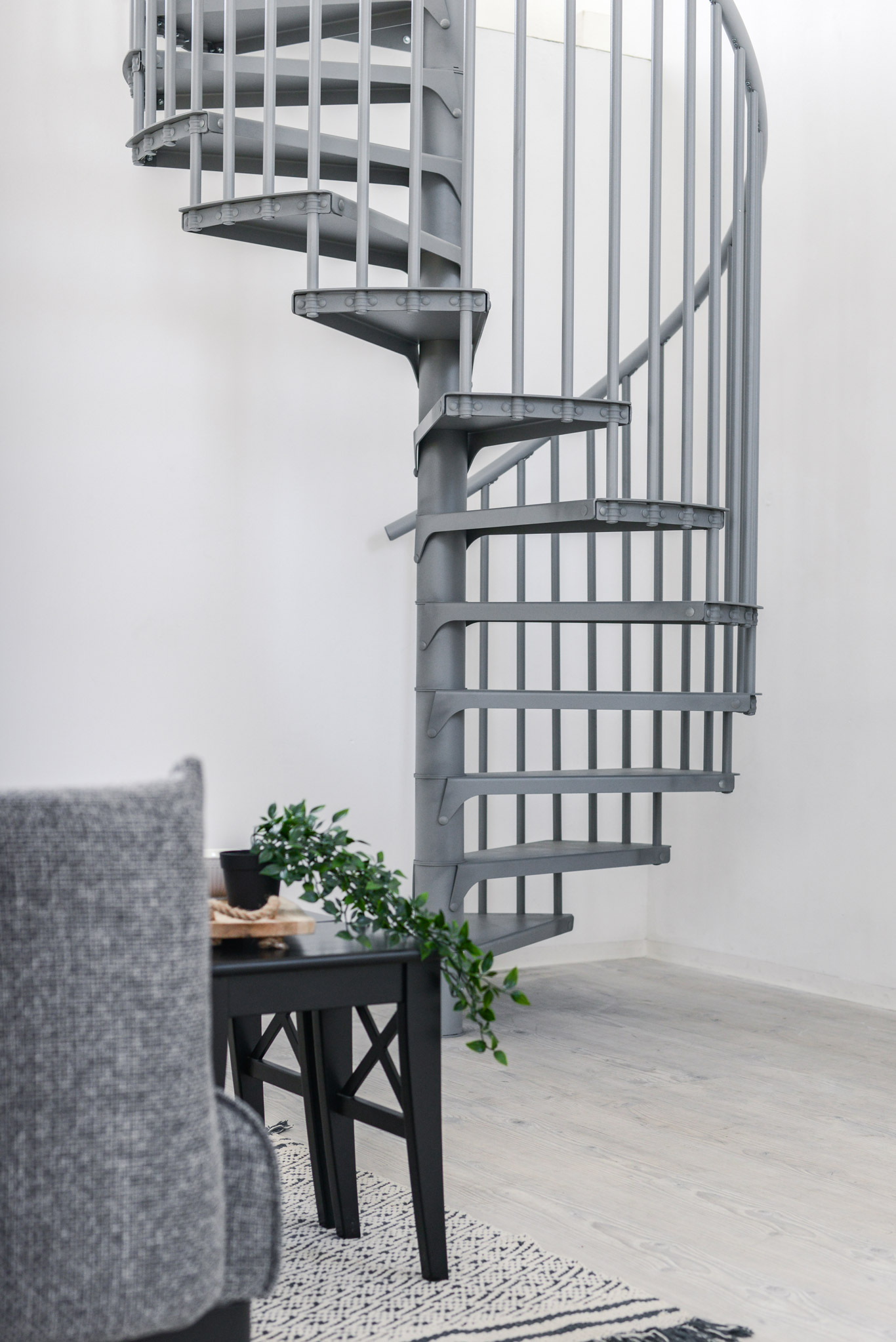 Die graue Spindeltreppe Rondo Smart aus Metall steht in einem modernen Wohnraum. Im Vordergrund ein schwarzer Tisch mit Deko und grauem Stuhl, im Hintergrund sind die filigranen Stufen und das senkrechte Geländer der Treppe sichtbar.