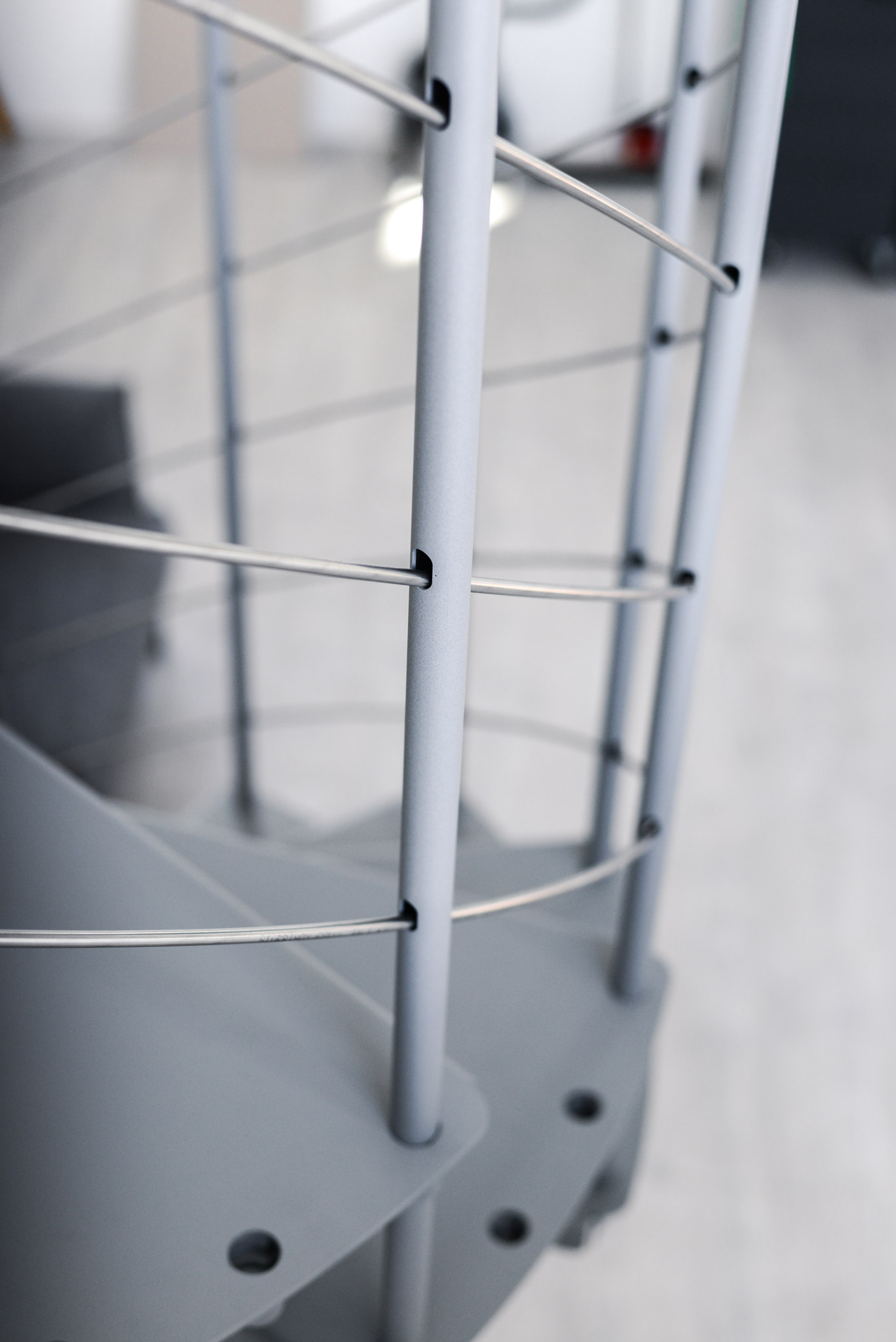 Nahaufnahme des Geländers der Spindeltreppe Spino Smart in Grau. Dünne Edelstahldrähte verlaufen waagerecht durch senkrechte Metallstäbe und bilden die Absturzsicherung entlang der gewendelten Metalltreppe.