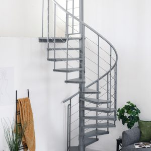 Spindeltreppe Spino Smart in Grau aus Metall im modernen Wohnraum. Die gewendelte Treppe führt nach oben, das Geländer besteht aus senkrechten Streben und Edelstahlseilen. Im Hintergrund stehen ein Sofa, Pflanzen und Deko.