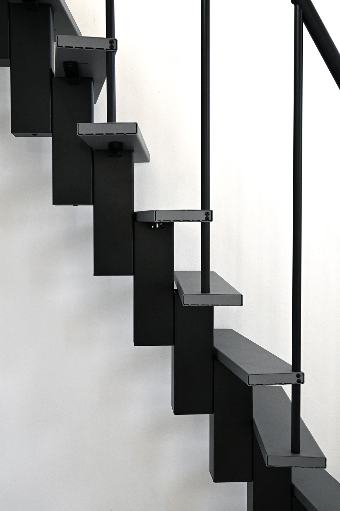 Seitliche Detailansicht einer modernen Raumspartreppe mit abwechselnd versetzten Metallstufen in Grau. Die Konstruktion ist schwarz, mit senkrechten Rundstäben als Geländer. Die ungeraden Stufen ermöglichen eine platzsparende und funktionale Nutzung bei begrenztem Raumangebot.