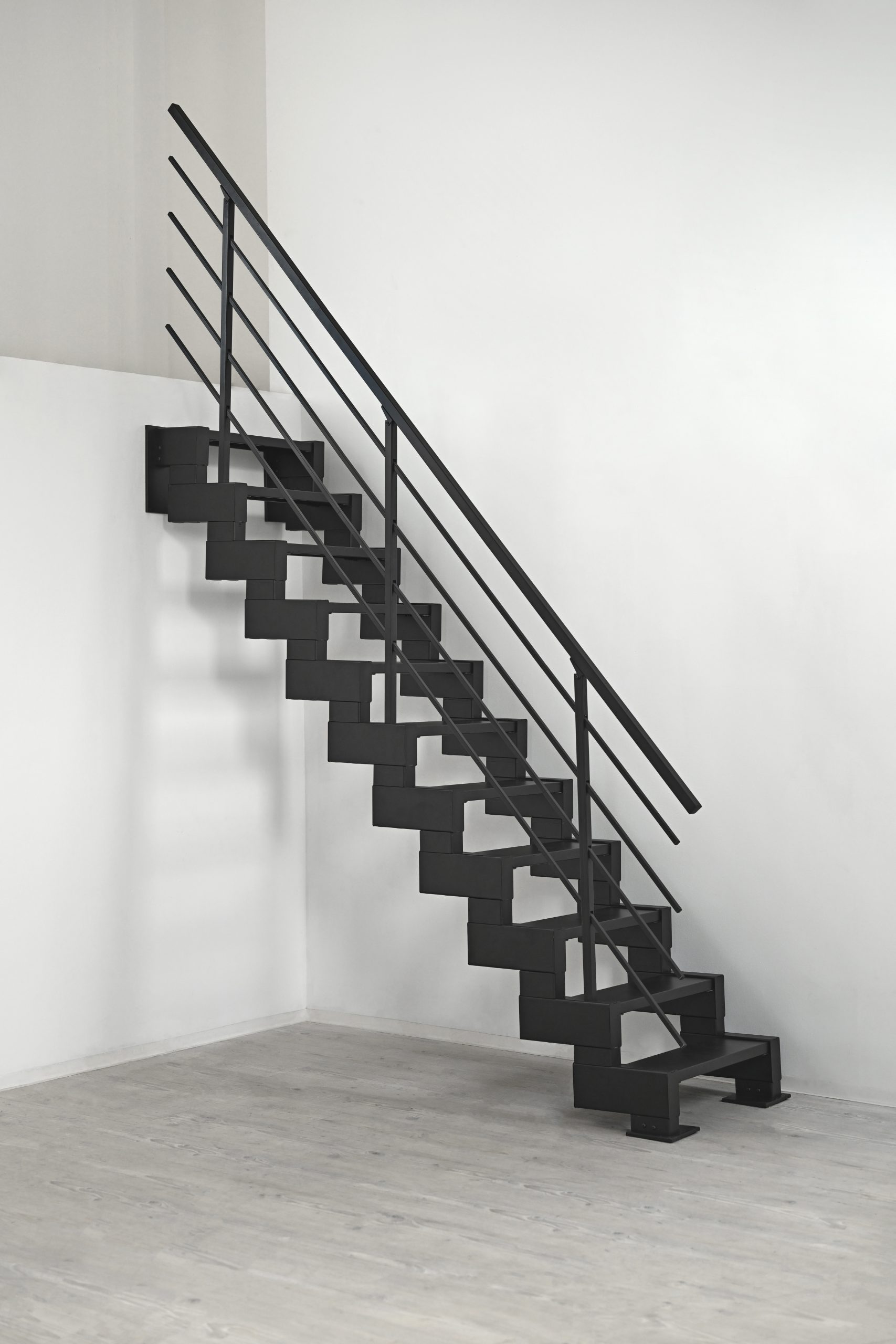 Geradläufige Raumspartreppe Modulario Mini in Schwarz mit Metallstufen. Die Treppe ist an einer weißen Wand montiert und steht auf einem hellen Holzfußboden. Das moderne Metallgeländer bietet sicheren Halt.