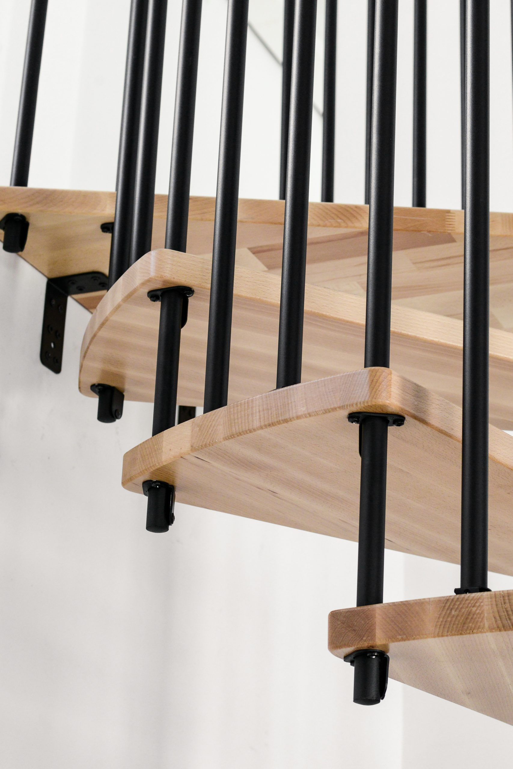 Nahaufnahme der Treppe Spiral Effect mit hellen Holzstufen und schwarzen Metallstäben. Die Stufen sind seitlich an der Wand verankert und verlaufen spiralförmig nach oben. Der Hintergrund ist schlicht weiß.