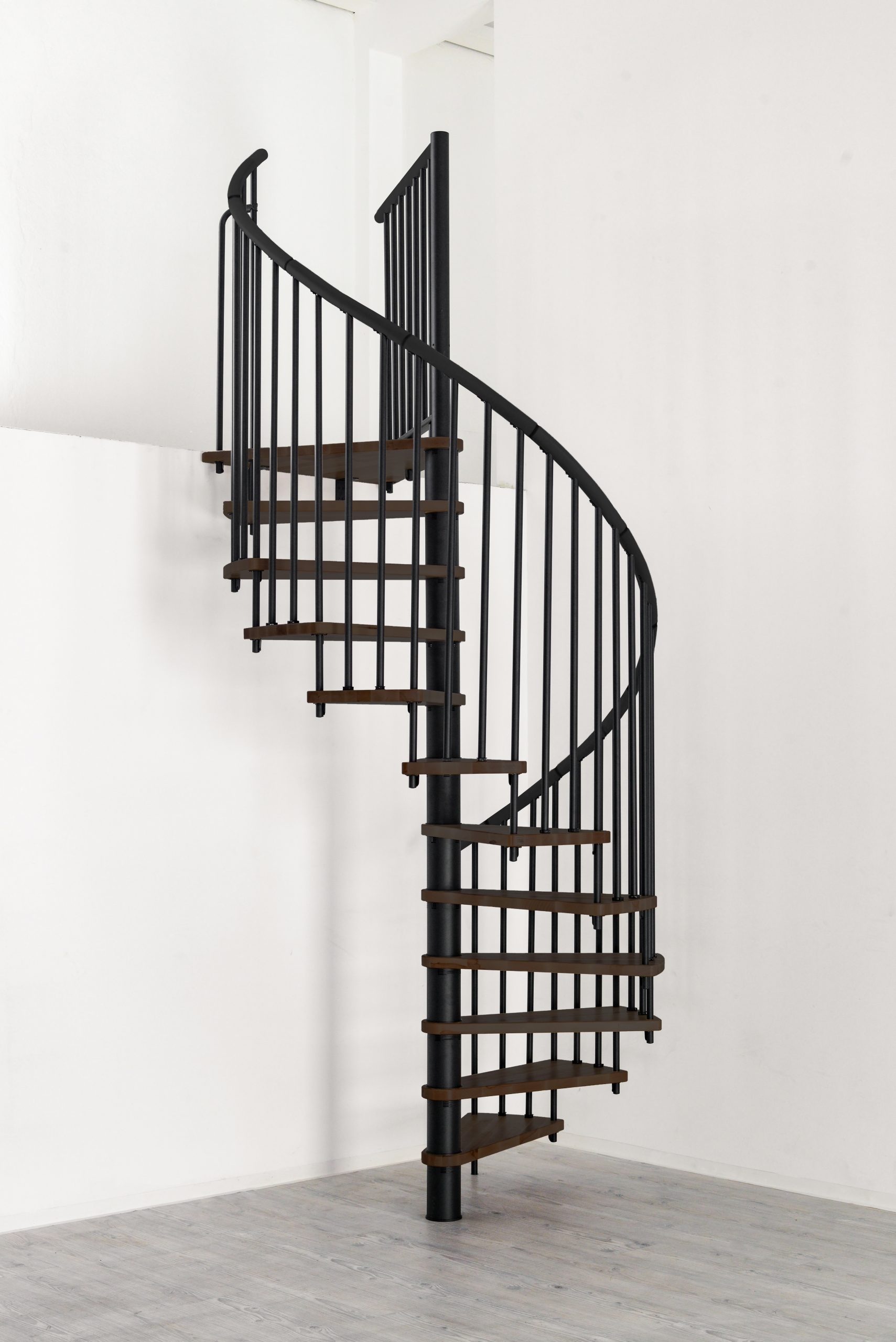 Spindeltreppe Spiral Wood mit dunklen Stufen in Walnussoptik und schwarzem Stahlgeländer mit Aluminium-Handlauf, aufgebaut in einem Raum mit grauem Boden und weißen Wänden.