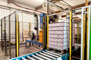 Automatisierte Verpackungsanlage mit einem Roboterarm, der Kartons auf einer Palette stapelt. Die Anlage ist von einem Sicherheitsgitter umgeben und steht in einer Produktionshalle.