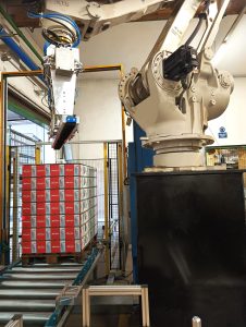 Ein Industrieroboter in einer Verpackungsanlage stapelt Kartons auf einer Palette. Die Kartons sind auf einem Förderband positioniert, das System ist durch Schutzgitter gesichert.