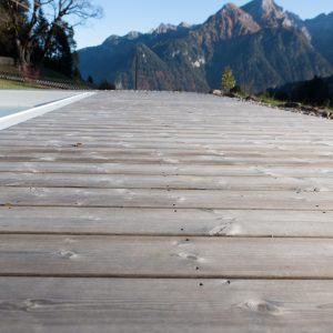 Holzterrasse aus Arum von Minka Wood, neben einem Pool gelegen, mit Blick auf ein traditionelles Berghaus und die umliegenden Berge unter blauem Himmel.