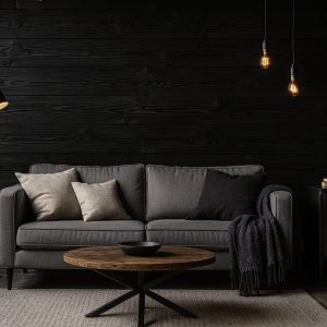 Dunkel gestalteter Wohnbereich mit grauem Sofa, runden Couchtisch aus Holz und Teppich, vor einer Wandverkleidung aus horizontal verlegten, schwarz gefärbten Holzbohlen, akzentuiert durch Stehleuchte, hängende Glühbirnen und dezente Dekoration.