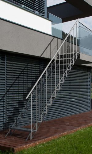 Außentreppe Sun Step aus verzinktem Stahl an einer modernen Hausfassade. Die Treppe hat 12 einzelne Stufen mit Gitterrost-Auflage, ein beidseitiges Geländer mit senkrechten Stäben und einem Handlauf aus Edelstahl. Zugang von der Terrasse in den Garten.