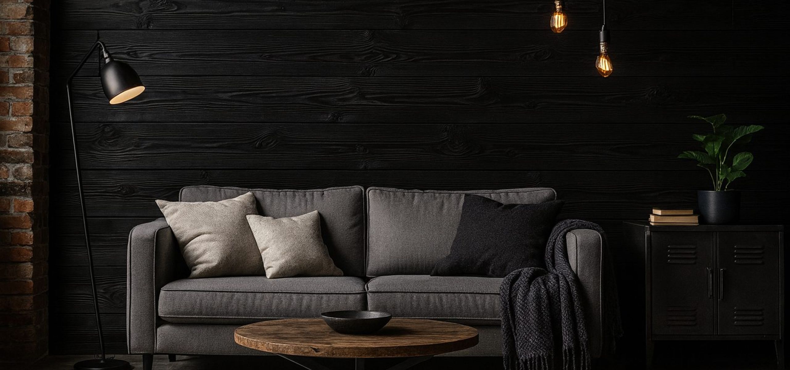 Dunkel gestalteter Wohnbereich mit grauem Sofa, runden Couchtisch aus Holz und Teppich, vor einer Wandverkleidung aus horizontal verlegten, schwarz gefärbten Holzbohlen, akzentuiert durch Stehleuchte, hängende Glühbirnen und dezente Dekoration.