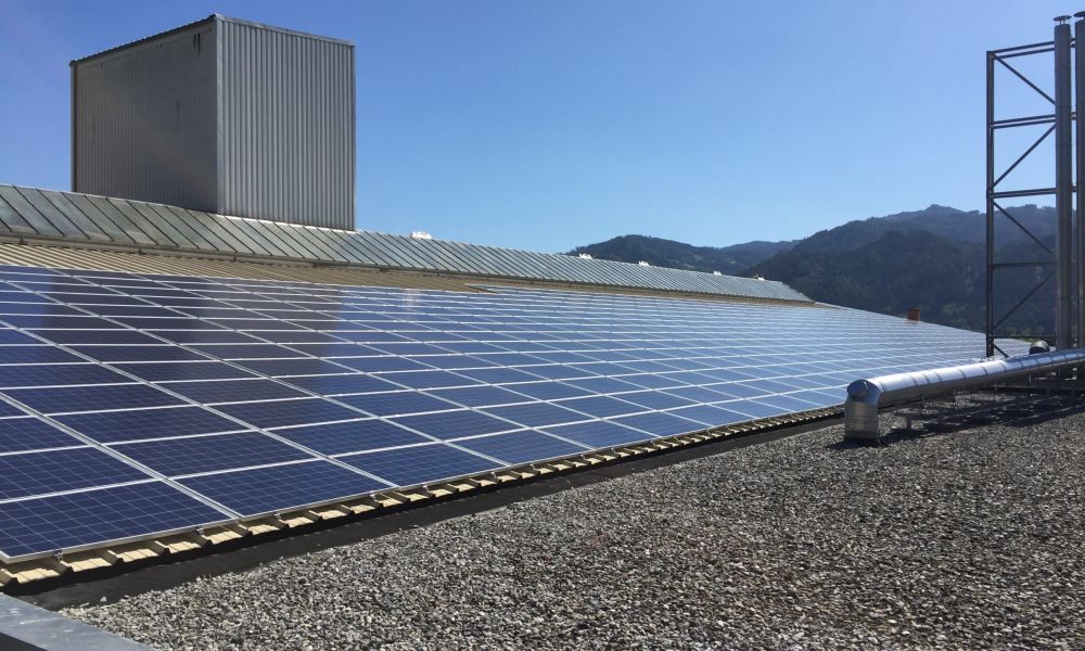 Photovoltaikanlage auf dem Flachdach eines Industriegebäudes bei sonnigem Wetter, im Hintergrund Berge und blauer Himmel.