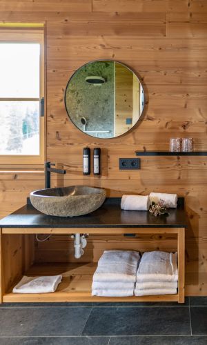 Waschtischbereich mit Naturstein-Waschbecken, Spiegel und Handtüchern. Die Wand ist mit gebürsteten Gladiola-Holzpaneelen gestaltet und sorgt zusammen mit den dunklen Steinfliesen für eine warme und moderne Atmosphäre im Almwelt Schwab-Walcher.