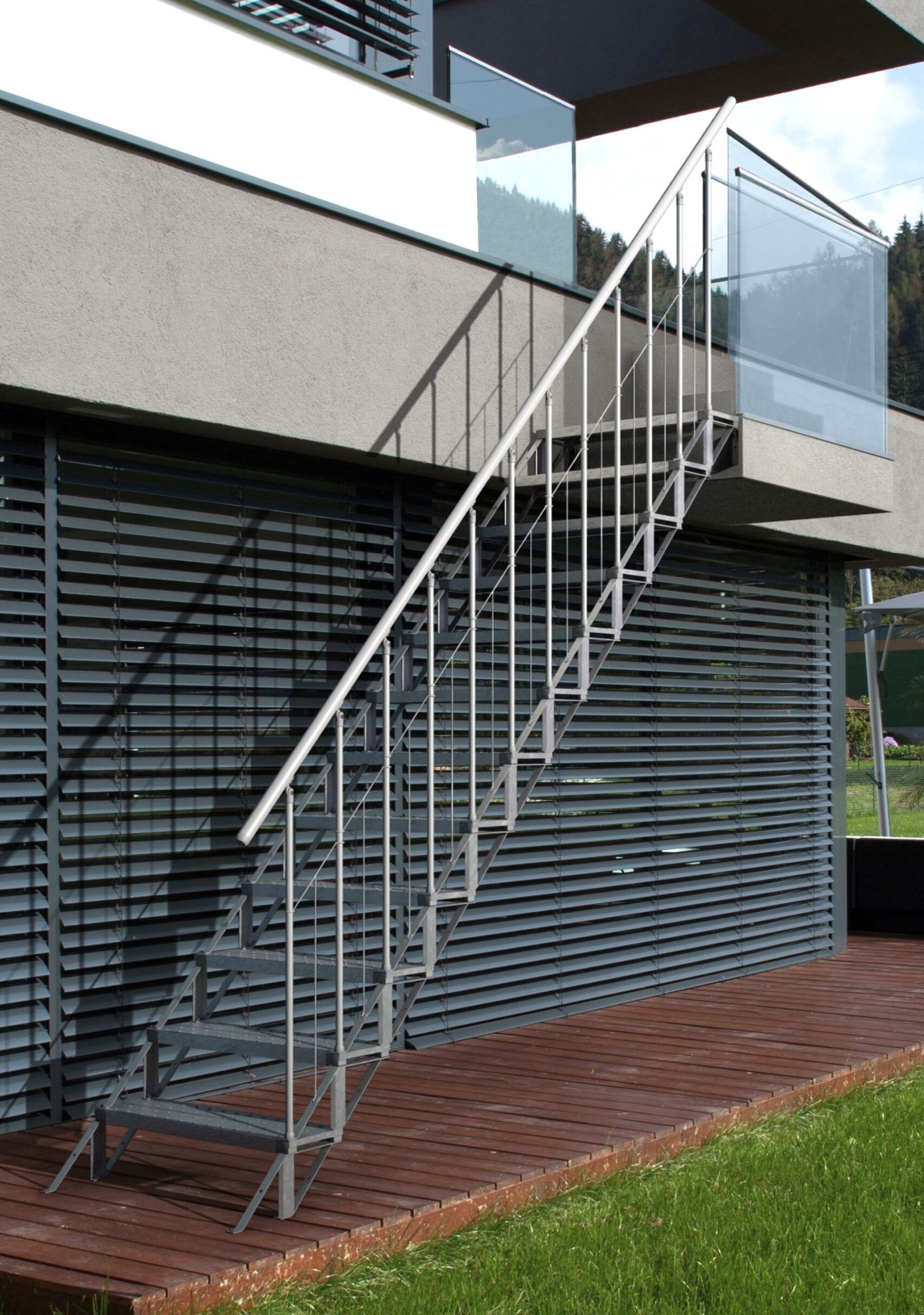 Moderne Außentreppe aus Metall mit Geländer, die von einer Holzterrasse zu einem Balkon mit Glasbrüstung führt.