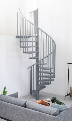 Graue Spindeltreppe Rondo Smart aus Metall mit runden Stufen und senkrechten Geländerstäben. Die platzsparende Treppe führt spiralförmig nach oben und steht in einem modernen Wohnraum mit Sofa, Pflanzen und Kleiderständer.