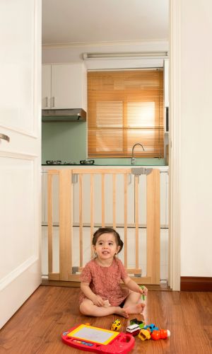 Kinderschutztür Britt in Türrahmen montiert, dahinter eine Küche. Ein kleines Kind spielt sicher im Vordergrund auf dem Holzboden mit buntem Spielzeug. Die Tür besteht aus hellem Holz mit vertikalen Streben und grauen Schließelementen.