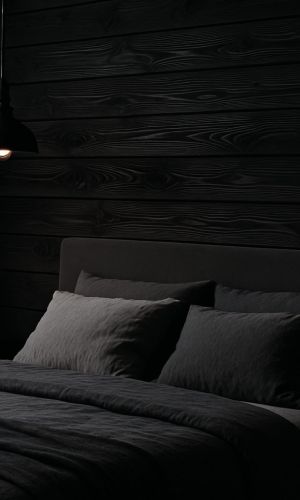 Dunkles, stimmungsvoll beleuchtetes Schlafzimmer mit Polsterbett und dunkler Bettwäsche, vor einer Wandverkleidung aus horizontal verlegtem, schwarz gefärbtem Holz Black Dahlia von Minka Wood, akzentuiert durch eine schwarze Pendelleuchte und dezente Nachttischbeleuchtung.